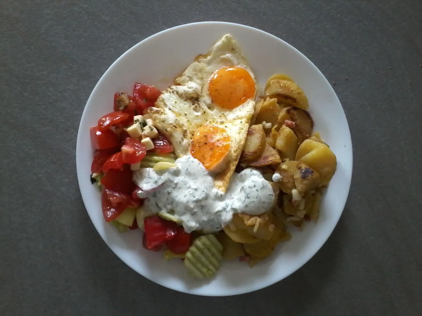 Teller mit Bratkartoffeln, zwei Spiegeleiern, Tomatensalat und einem Klecks Kräuterquark.