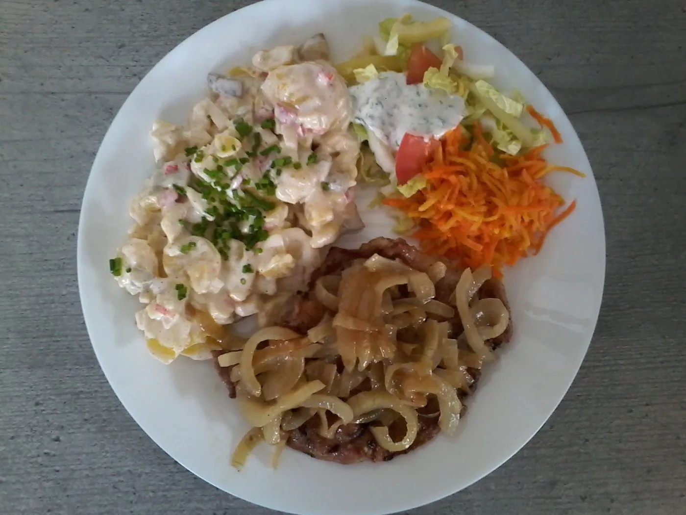 Rostbrätl mit Röstzwiebeln, Kartoffelsalat, Karottensalat und gemischtem Salat auf einem Teller.